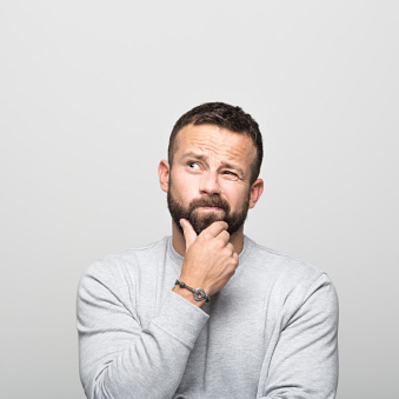 Portrait of worried bearded young man looking up with hand on chin