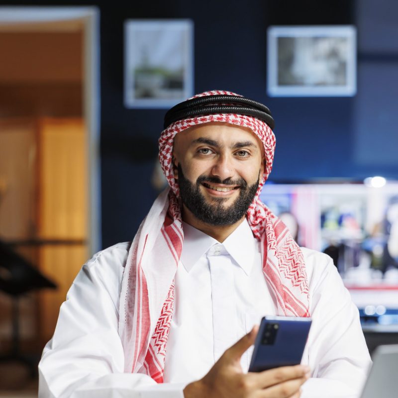 Male Arab person in profile, grinning at camera with his personal computer close by. Using contemporary technology, smiling Muslim guy with a cell phone, portrayed in portrait with laptop on table.