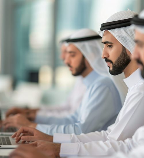 A row of men wearing traditional white robes work on their laptops in a modern office setting.